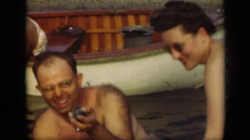 1948 Long Island home-movie frame showing a smiling man holding a small object beside a woman and canoe.