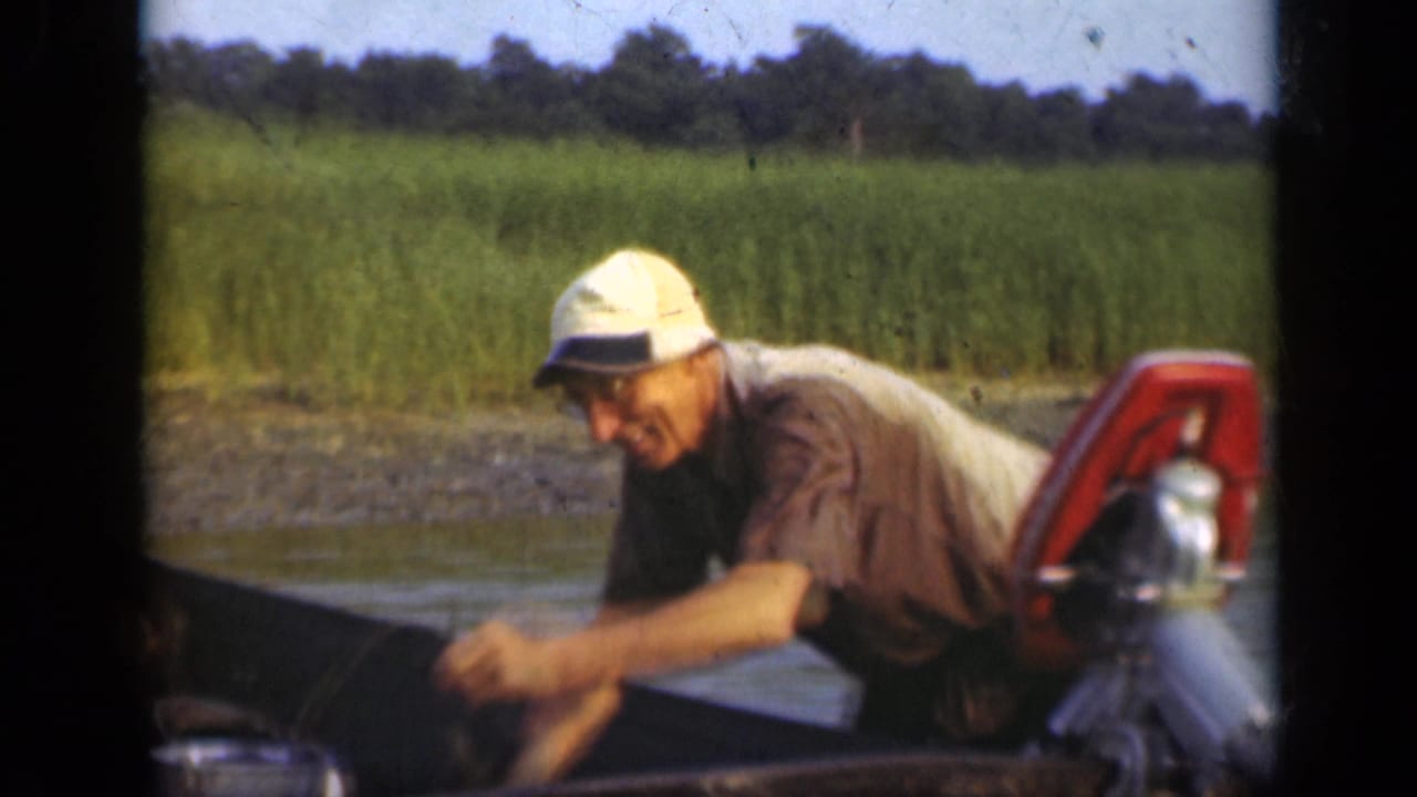 Man in a white cap leans over a small boat beside a red outboard motor on calm water.