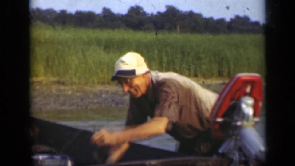 In 1948 Long Island home-movie footage, a fisherman leans over a boat near a red outboard motor with marsh grass behind him.