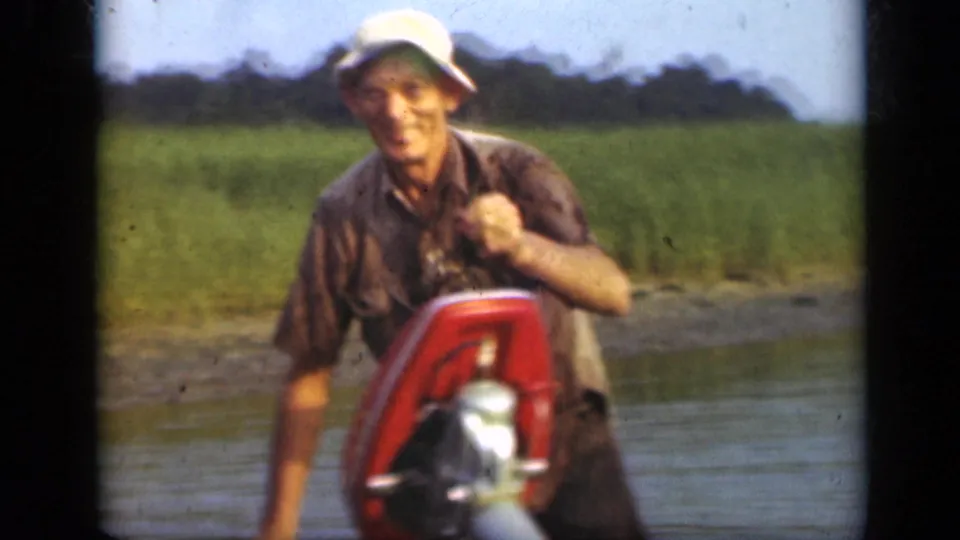 The Long Island fisherman smiles toward the camera as he lifts a dark fish beside the small boat and red motor.