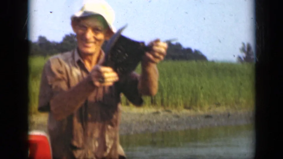 In a soft 8mm frame, the fisherman holds the catch upright for the camera against Long Island marshland and calm water.