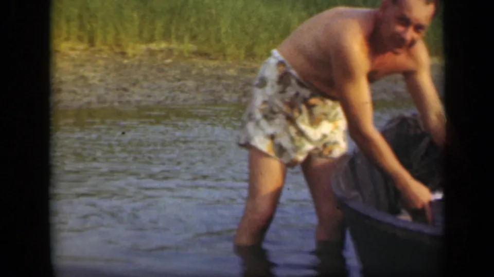 A shirtless man in patterned trunks bends beside a small boat in shallow Long Island water, handling something near a bucket or boat edge.
