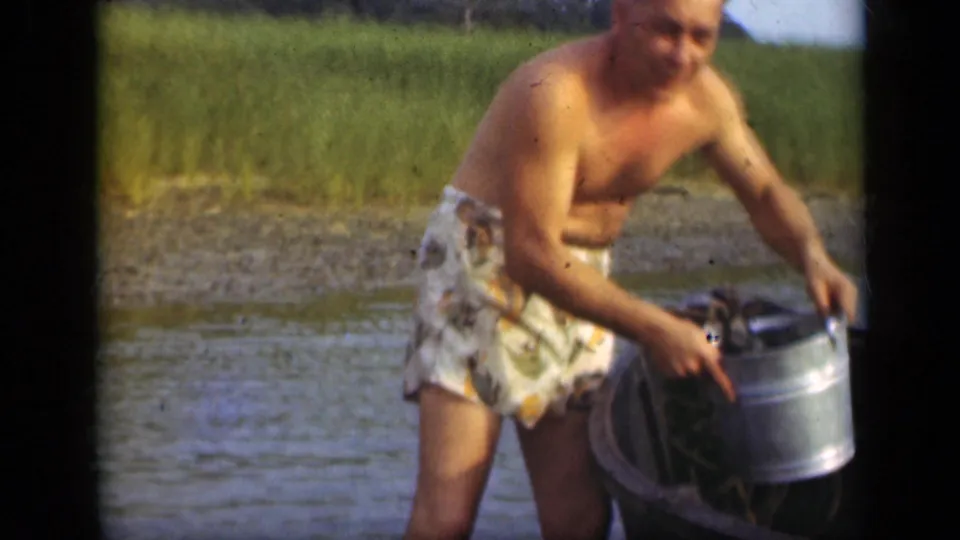 A shirtless man in patterned swim trunks handles a metal bucket beside a small boat in shallow water on Long Island.