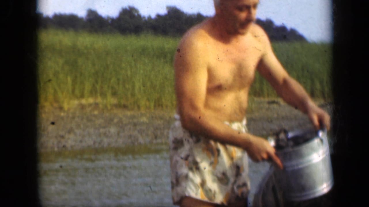 1948 Long Island home-movie frame showing a shirtless man holding a metal pail near shallow water with green reeds behind him.