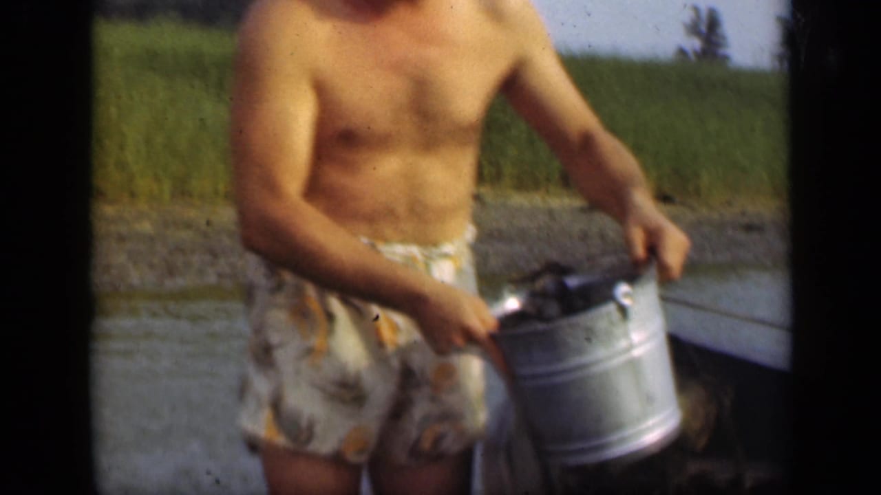 Man carries a metal bucket near a boat edge with reeds and water in the background.