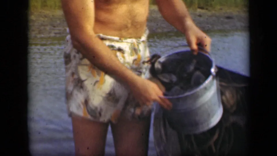 Close 8mm frame of the man's torso and metal bucket, with dark shellfish visible inside near the waterside boat.