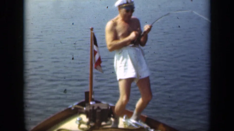 Man in white shorts stands on a Long Island boat in 1948, holding a fishing pole near a small flag and calm water.