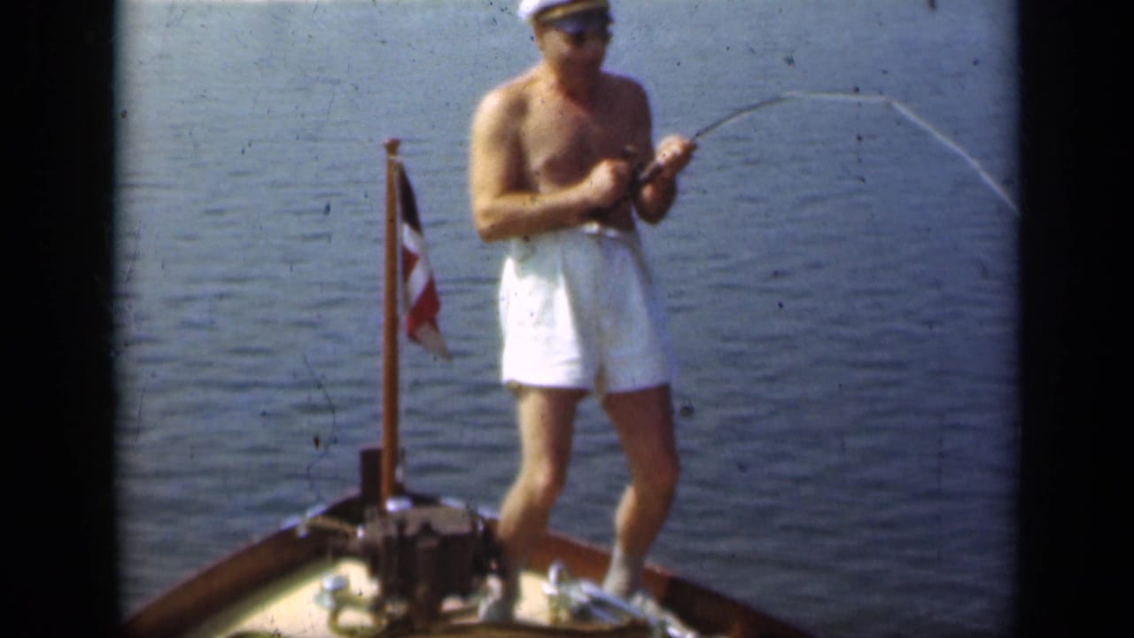 Shirtless man in white trunks stands on a boat bow holding a fishing rod over calm water.