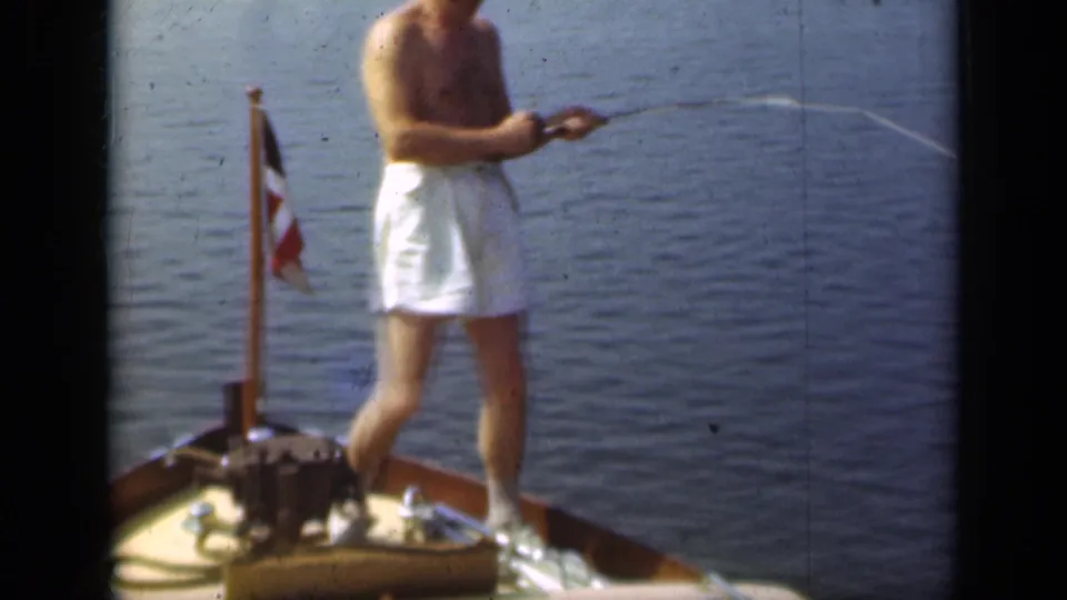 The man stands near the small flag on the wooden bow, pulling or casting a fishing line in the 1948 Long Island footage.