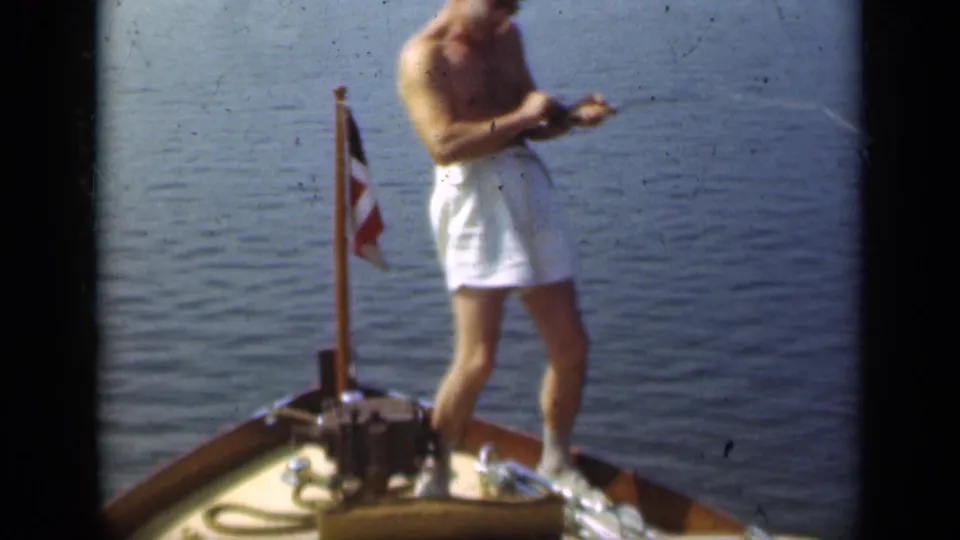 A shirtless adult man in white trunks leans back on the wooden bow as he works the fishing rod over calm Long Island water.
