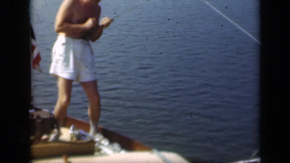 A shirtless man in white swim trunks appears to reel a line from the bow of a wooden boat on Long Island in 1948.