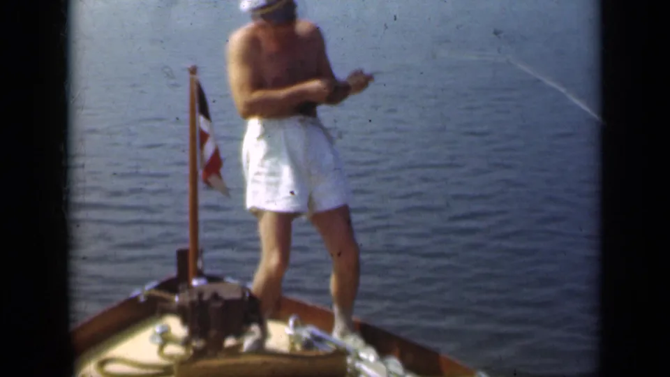 A shirtless man in white trunks and a captain-style hat reels from the bow of a small boat, with water and a flag behind him.