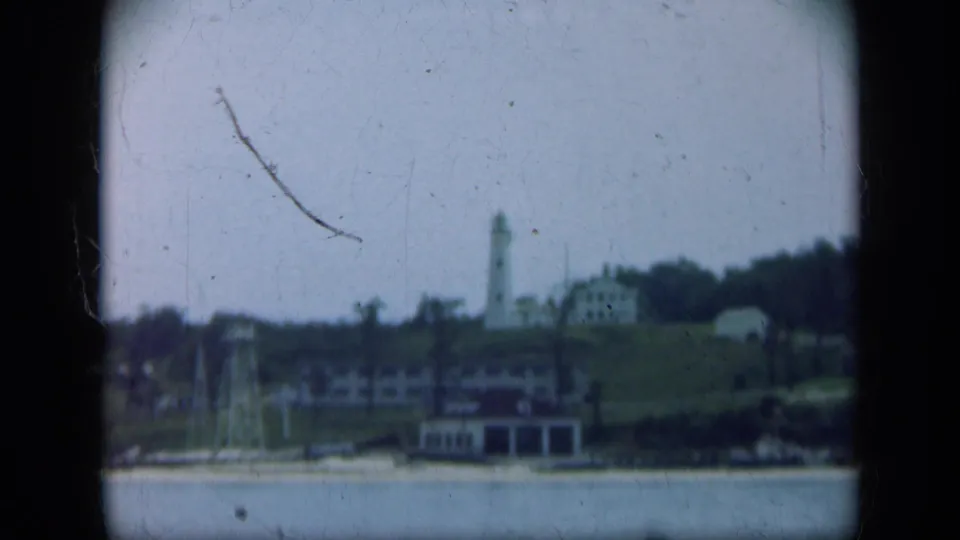 Distant Long Island shoreline in 1948 with water, low buildings, trees, and a lighthouse-like tower under hazy exposure.