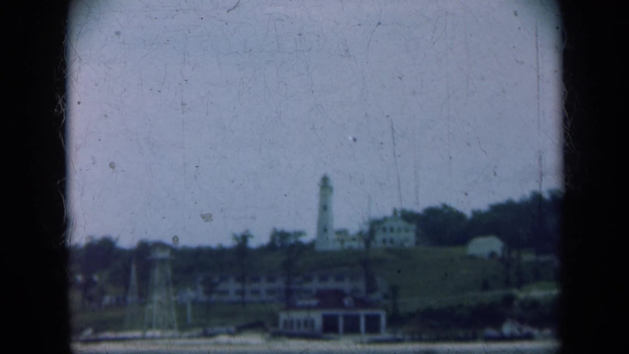 Distant lighthouse and shoreline buildings seen across calm water through dark 8mm frame edges.