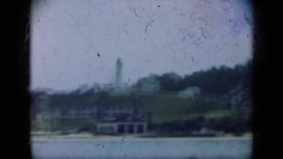 Distant Long Island lighthouse on a grassy rise, with trees and low waterfront buildings below in hazy 8mm color.