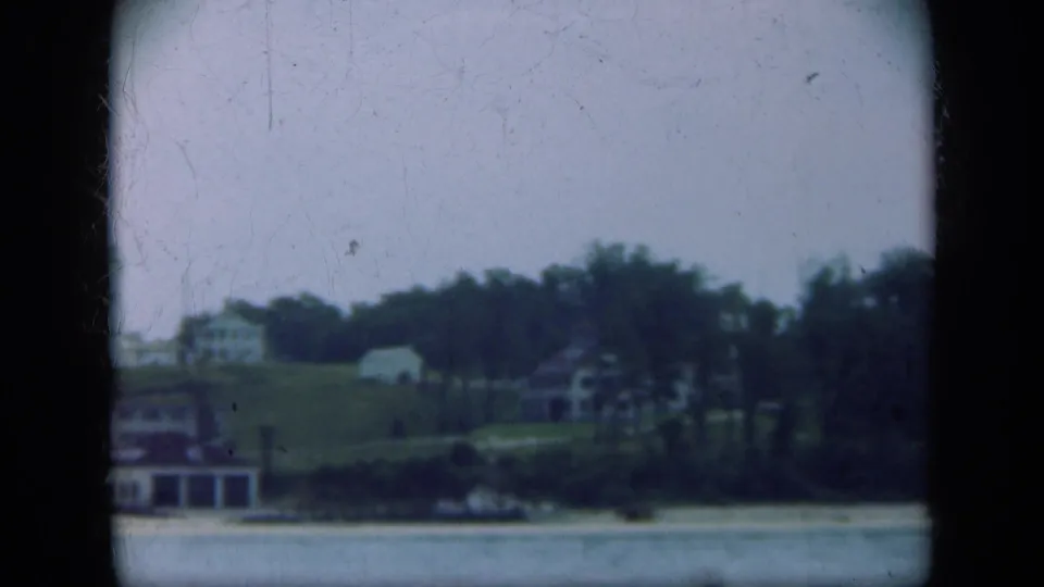 1948 Long Island shoreline view with low marina buildings, trees, a grassy hill, and calm water, framed by 8mm gate edges.