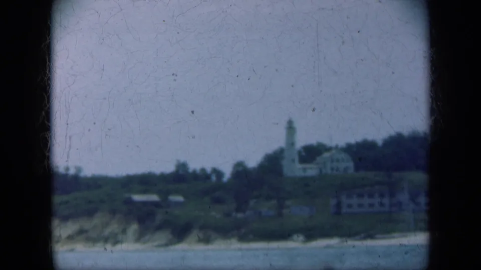 Long Island coastal frame showing a white lighthouse near houses and trees above a shoreline slope, with water in the foreground.