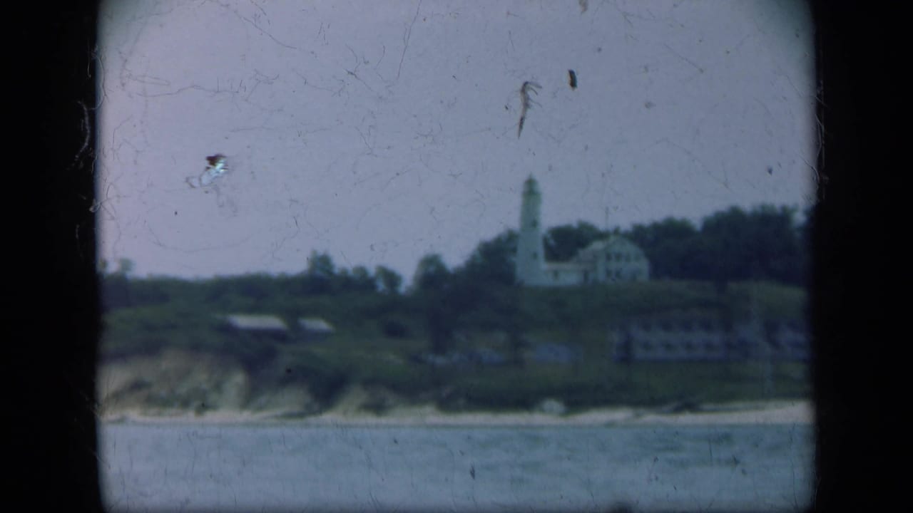 1948 Long Island view of a white lighthouse and low buildings on a grassy coastal hillside beyond calm water.