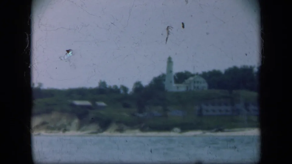1948 Long Island view of a white lighthouse and low buildings on a grassy coastal hillside beyond calm water.