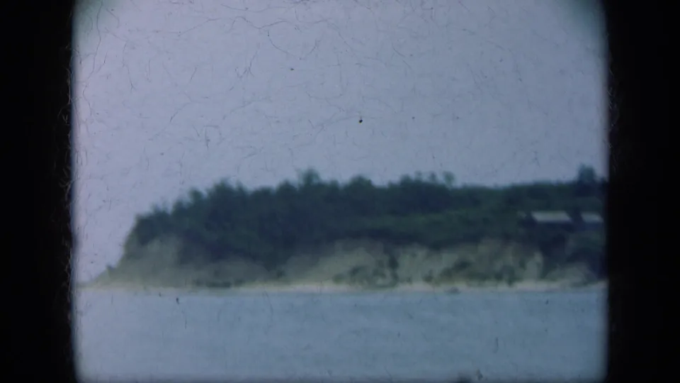 Soft 8mm frame of the Long Island shoreline, showing water, an eroded bluff, trees, and distant hillside structures.