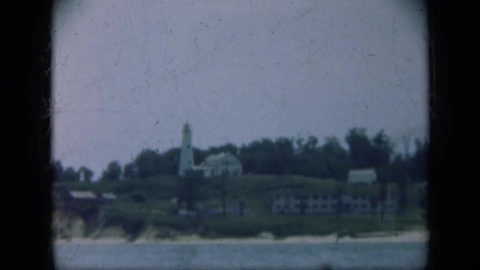 1948 Long Island home-movie frame with a lighthouse, clustered hillside buildings, trees, sandy shore, and water.