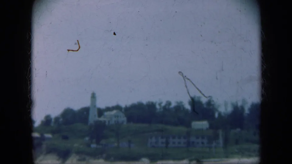Distant Long Island lighthouse scene on faded 8mm film, with houses on a bluff and beach below.