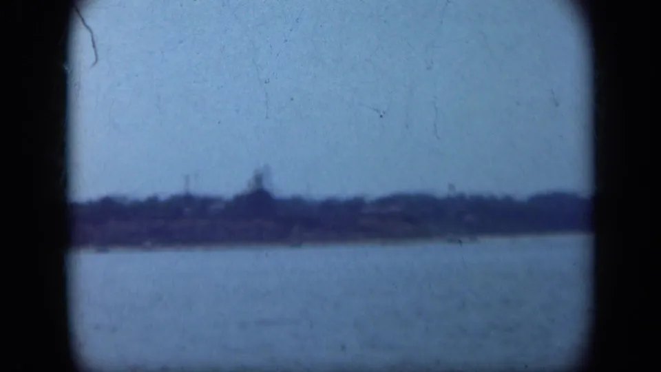 1948 Long Island shoreline seen from a boat, with low buildings or trees along the water beneath a pale blue sky.