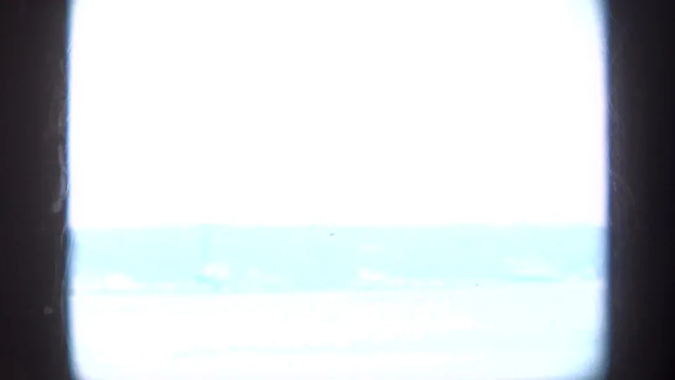 Window-framed coastal view in the Long Island clip, with pale water, hazy sky, and distant low land.