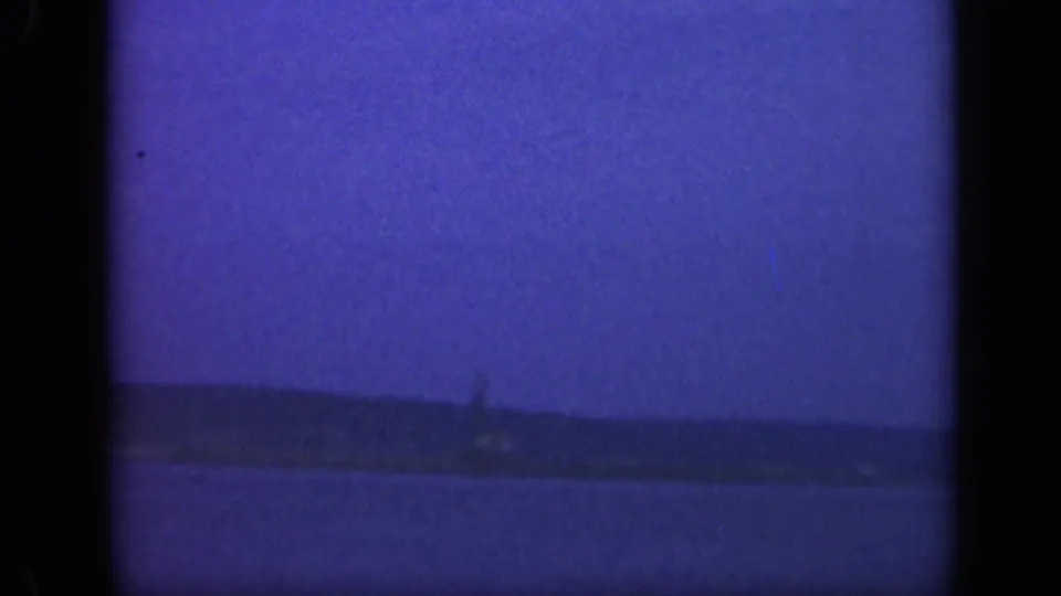 Blue-tinted 8mm view of water and a low Long Island shoreline, with a faint distant tower or lighthouse form near the horizon.