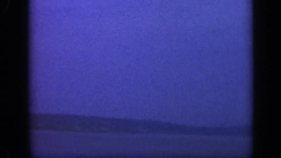 1948 Long Island coastal frame with a dark low shoreline, calm water, and blue-tinted 8mm grain as the view fades darker.