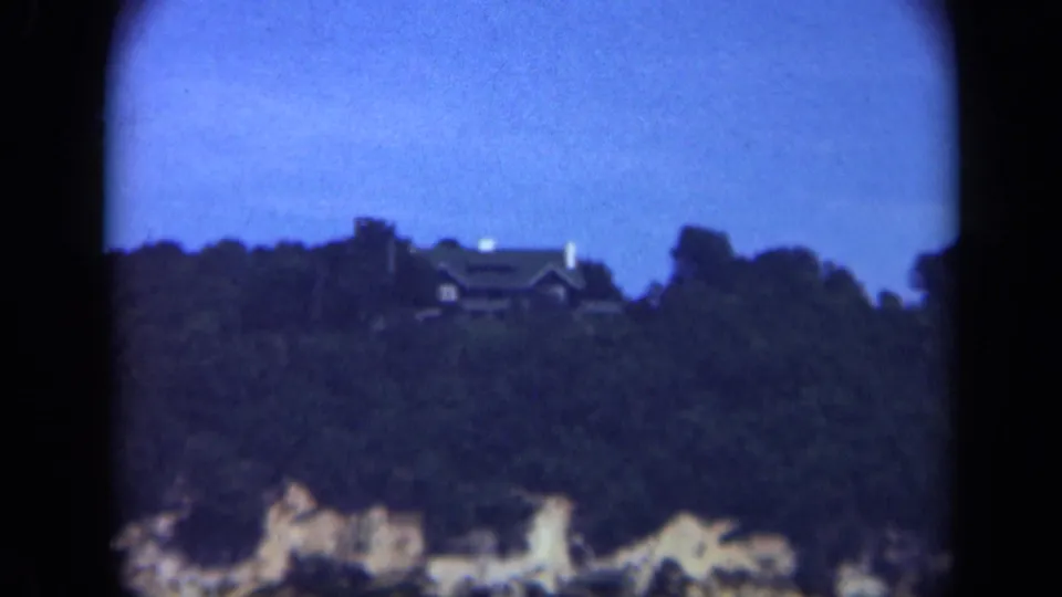 A distant Long Island house sits among dark trees on a cliff above the shoreline in a soft 8mm frame.