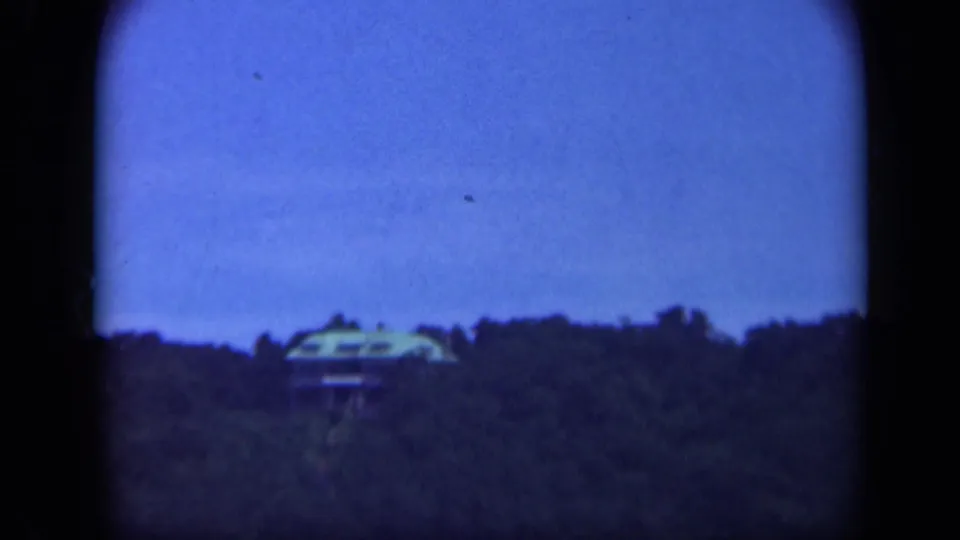 1948 Long Island frame with a distant house partly obscured by trees on a wooded ridge beneath a clear blue sky.