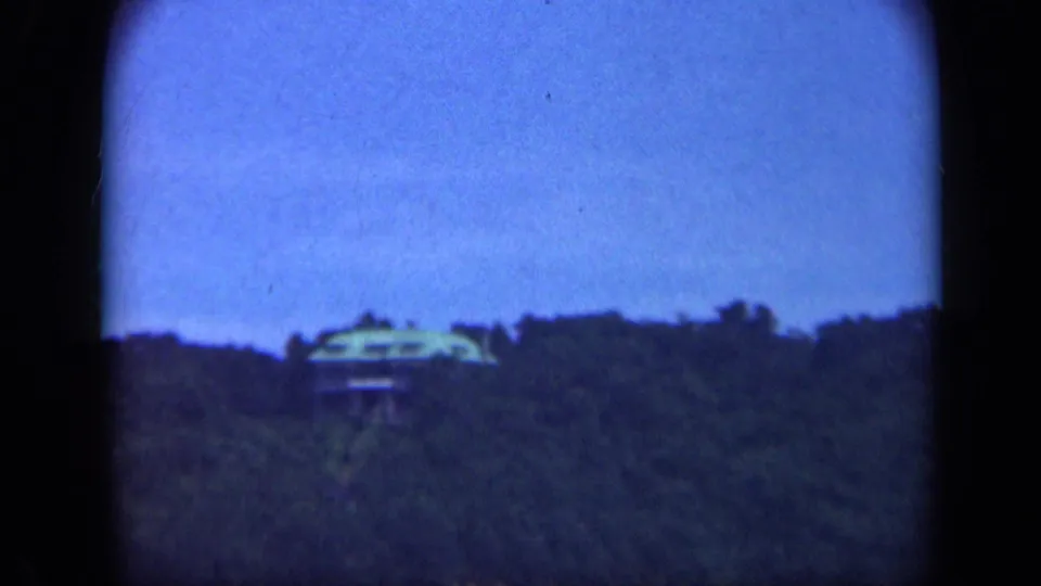 Mid-century 8mm Long Island view centering a distant hilltop house above dense trees, with soft focus and blue color shift.