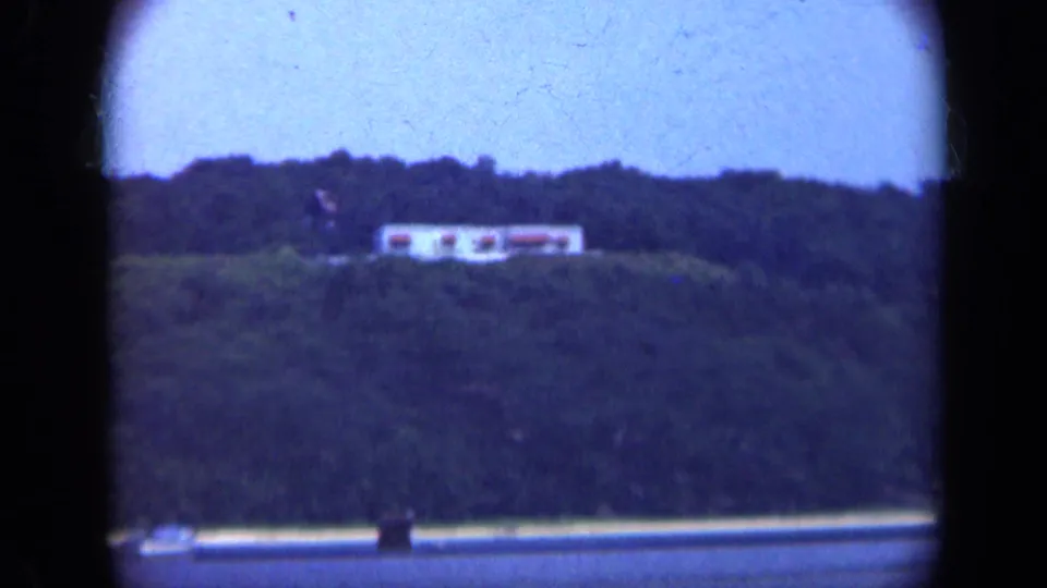 1948 Long Island 8mm frame showing a distant pale structure on a wooded hillside above a strip of water.