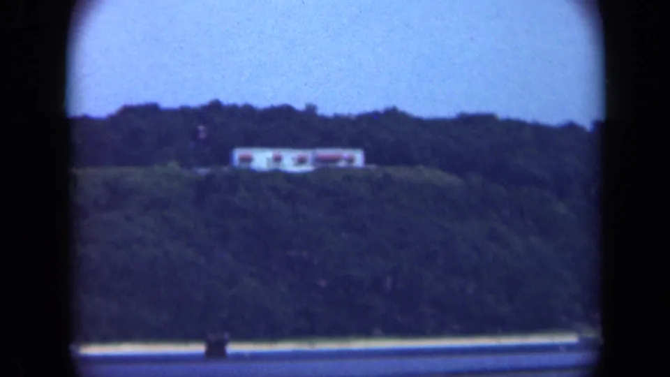 1948 Long Island home-movie frame with a distant hillside structure, dark trees, water foreground, and blue dusk sky.