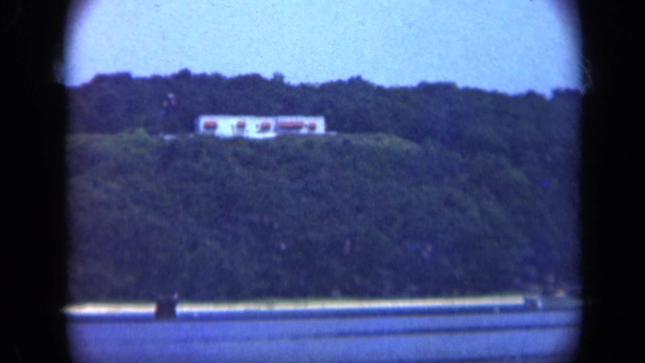 1948 Long Island 8mm view from the water toward a white hilltop building above a wooded shoreline.