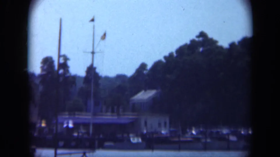1948 Long Island marina view with docked boats, a tall mast, and flags beside a wooded waterfront.