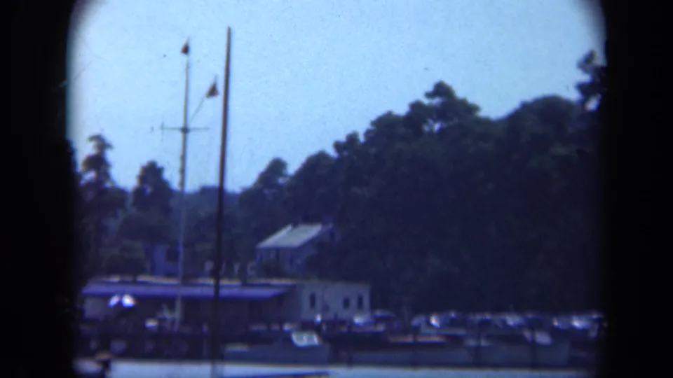 Long Island waterfront marina in 1948 with a dock house, tall mast, flags, parked cars, and dense trees beyond the shore.