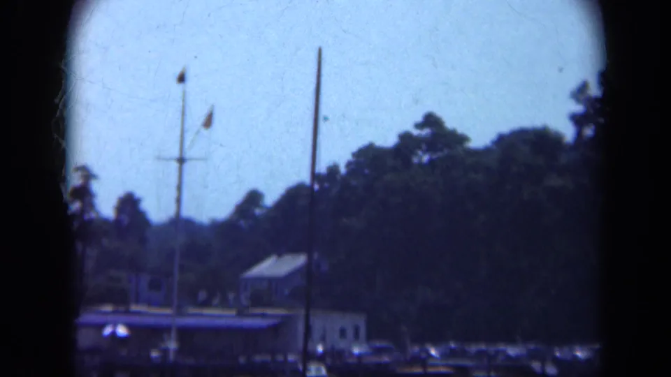 Boat-shot 1948 Long Island marina frame with mast and flags over a dock house, shoreline trees, parked cars, and water.