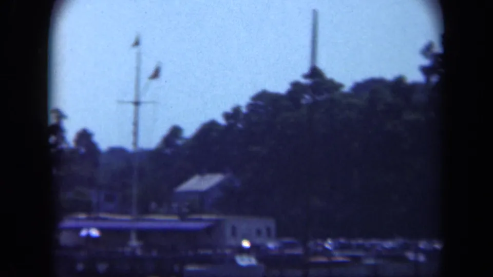 1948 Long Island marina scene from the water, with a dock house, mast, flags, parked cars, trees, and visible 8mm gate edges.