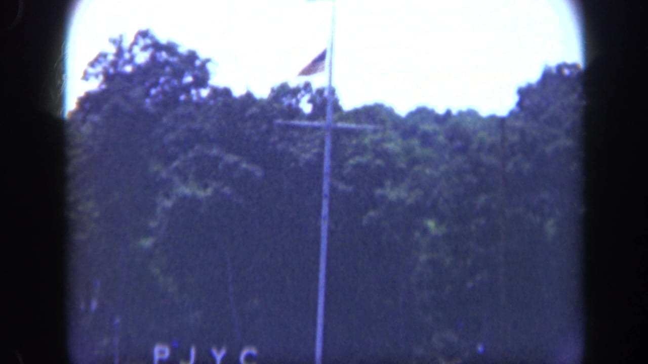 Flagpole and PJYC lettering seen through dark film gate edges above a wooded shoreline.