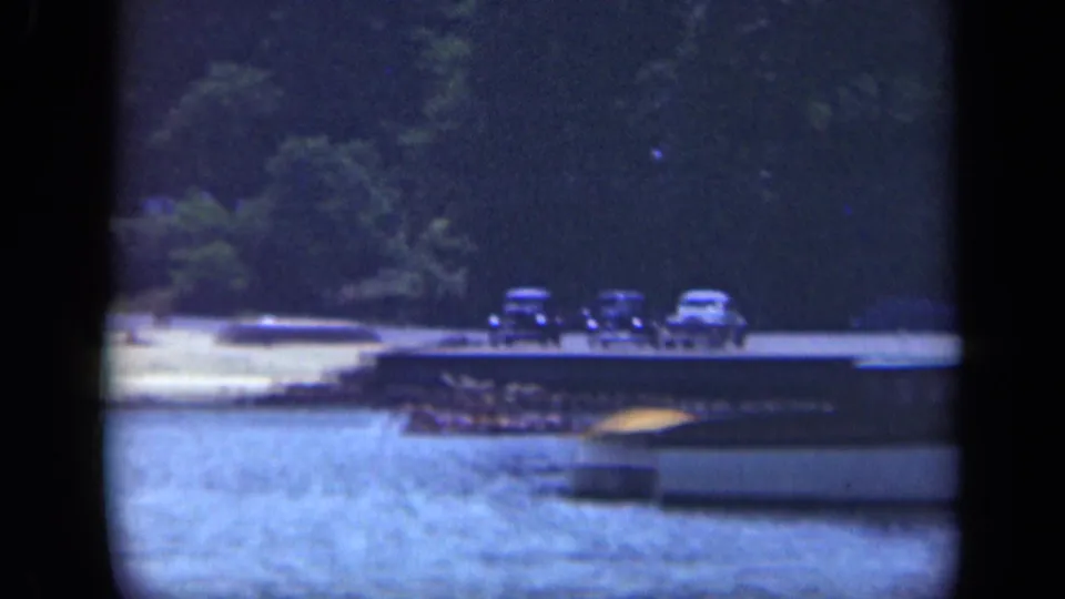 Classic cars sit near a Long Island waterfront dock, with water in the foreground and wooded shore behind.