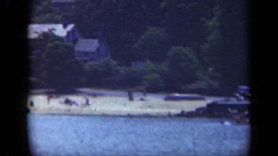 Long Island camp shoreline with a sandy beach, small structures, trees, water, and tiny figures near the shore.