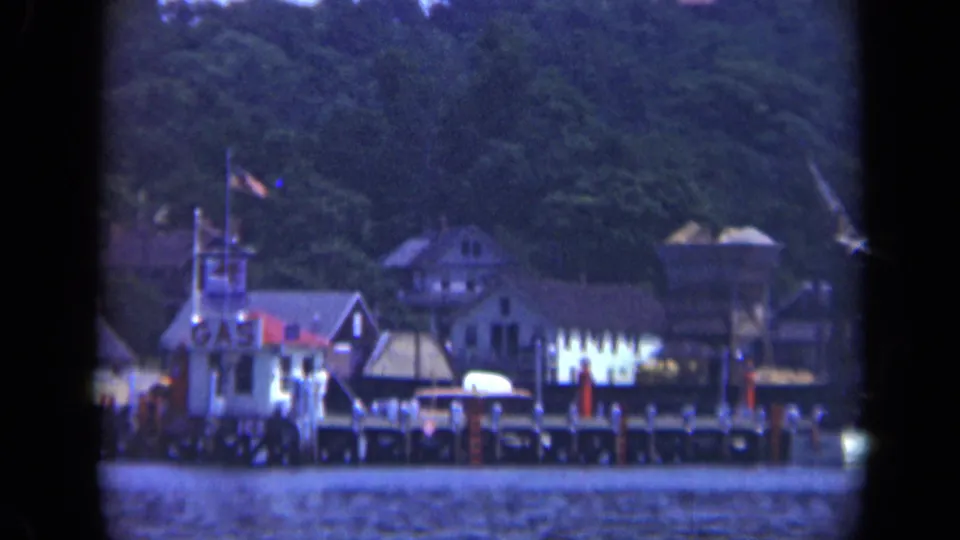 1948 Long Island waterfront camp or club area with flags, dock structures, parked cars, and people gathered near the buildings.