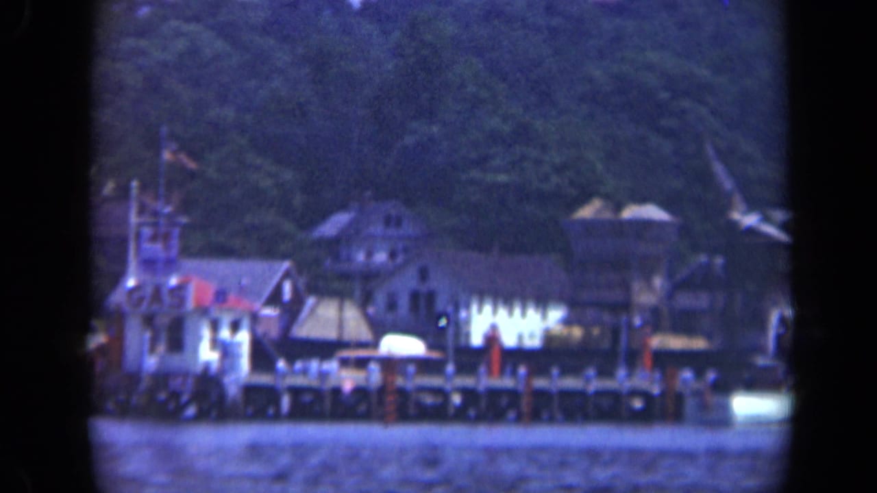 1948 8mm view of a Long Island waterfront with clustered buildings, dock pilings, boats, and trees in the background.
