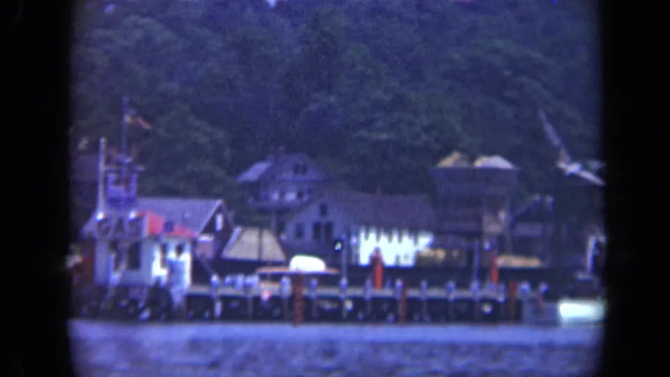1948 8mm view of a Long Island waterfront with clustered buildings, dock pilings, boats, and trees in the background.