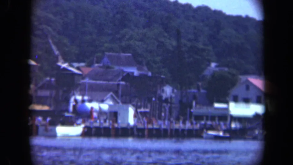 1948 8mm frame of a Long Island riverside town with a white shoreline building, boats, piers, and trees.