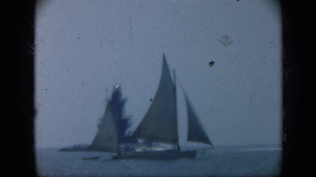 Large sailboat and smaller sailboat pass in front of a distant lighthouse on open water.