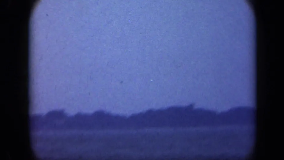 Blue-tinted 8mm frame from Long Island showing water in the foreground and a soft wooded horizon.