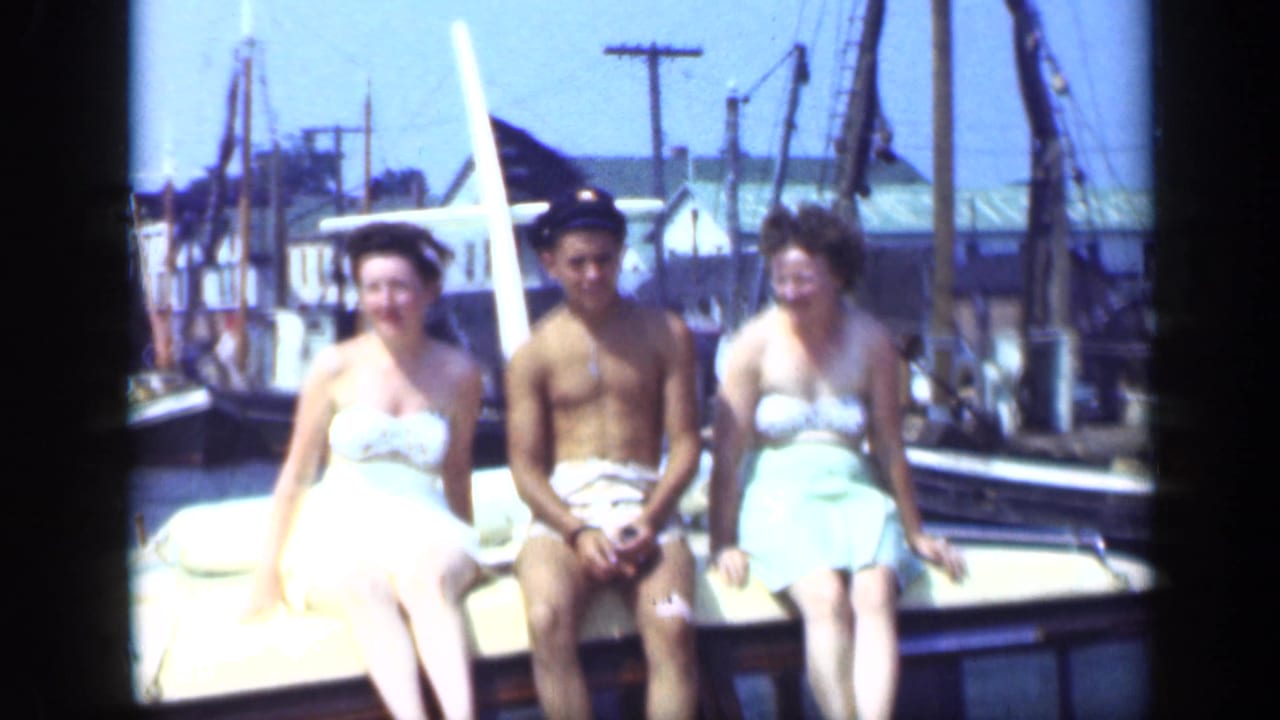 In 1948 Long Island home-movie footage, two women and a young man in a captain hat sit together on a boat at a marina.